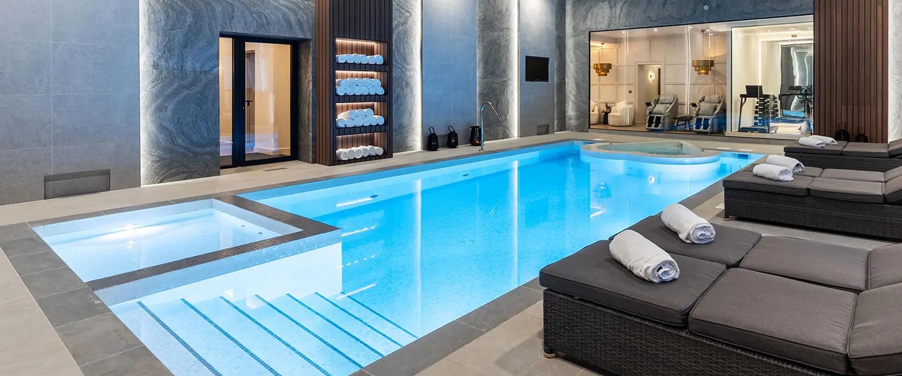 Indoor swimming pool with hot tub, poolside loungers with rolled towels, and a gym visible through a mirror in a luxury holi…