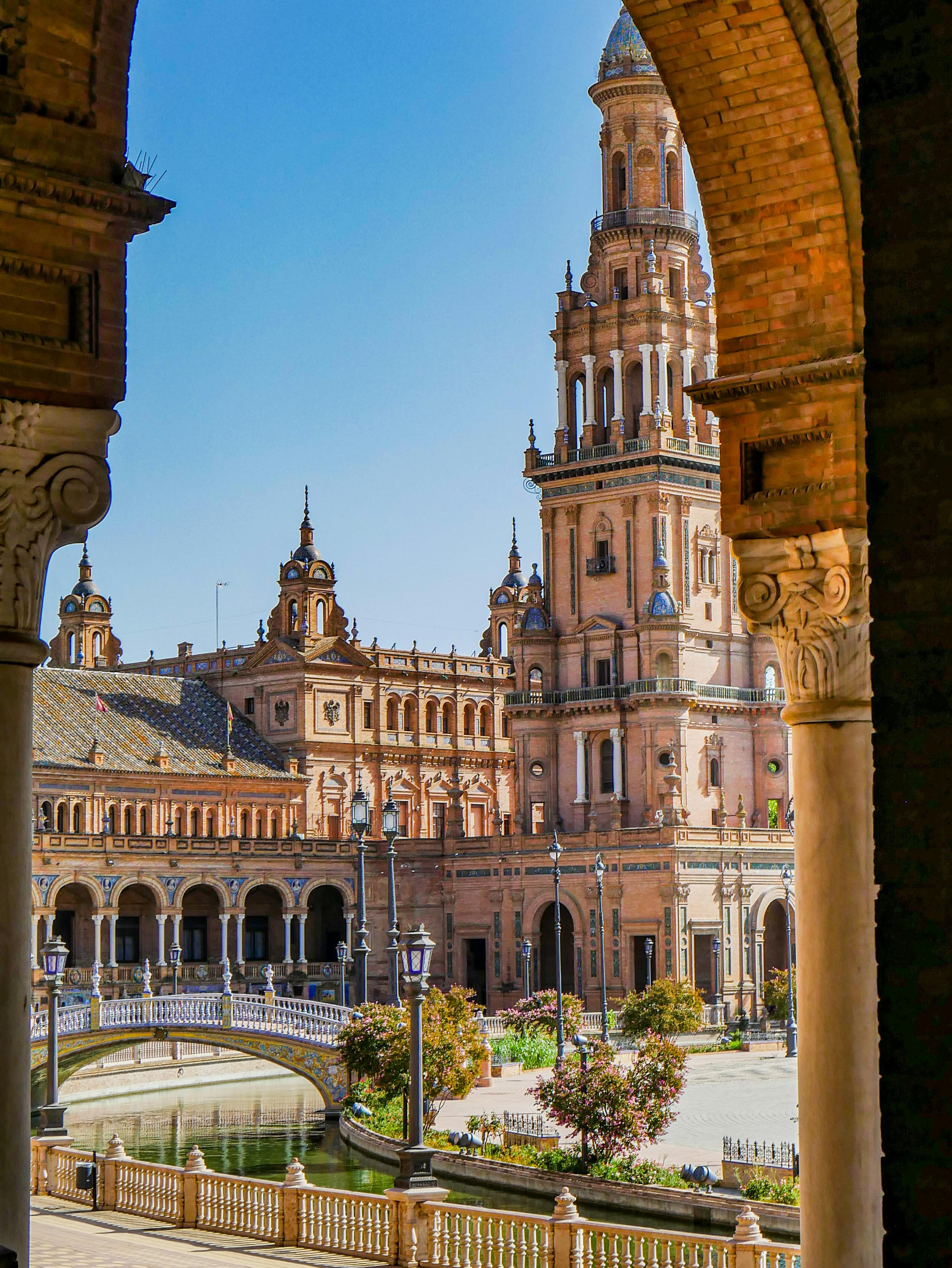 Plaza de España in Seville framed by a brick arch, showing the ornate Renaissance tower, tiled bridge, and canal under a cle…