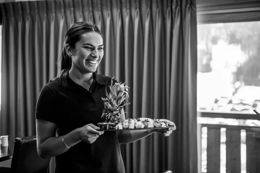 Smiling chalet staff member presenting welcome refreshments including fresh pineapple and treats on elegant serving tray