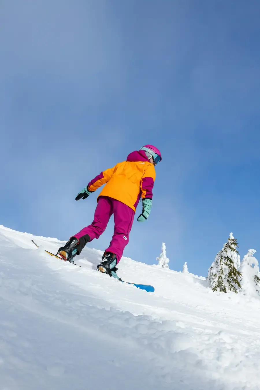 Child snowboarding down pristine alpine slope in bright orange and pink ski gear under clear blue sky