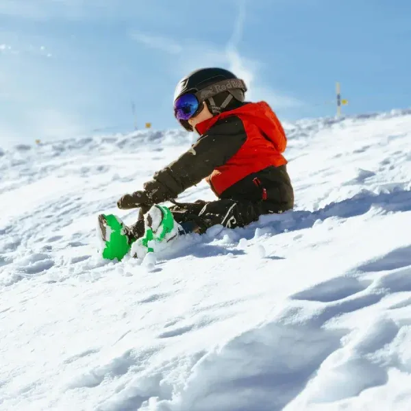 Child skiing down snowy mountain slope wearing red jacket and goggles with green skis on sunny winter day