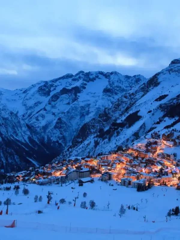 Alpine ski resort village illuminated at dusk beneath snow-covered French Alps peaks with ski slopes in foreground