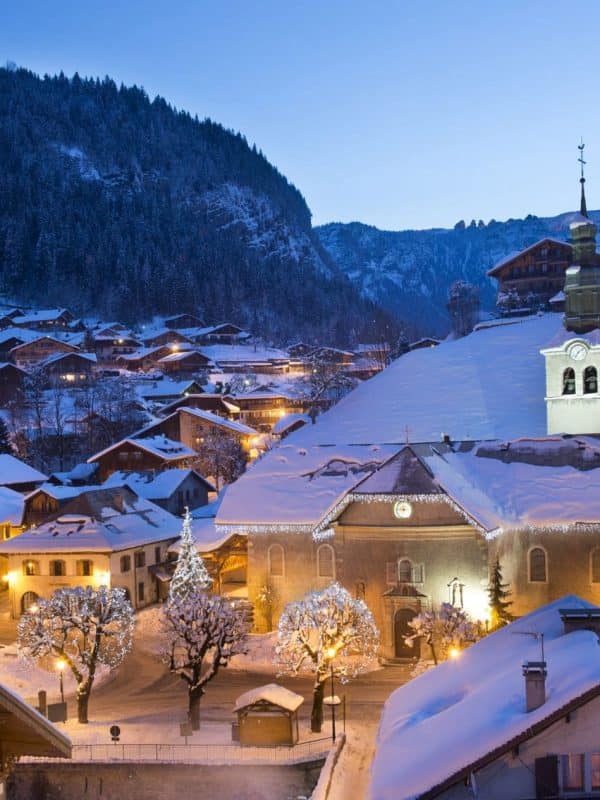 Snow-covered Alpine village at dusk with illuminated chalets, church spire and Christmas lights nestled in forested mountains