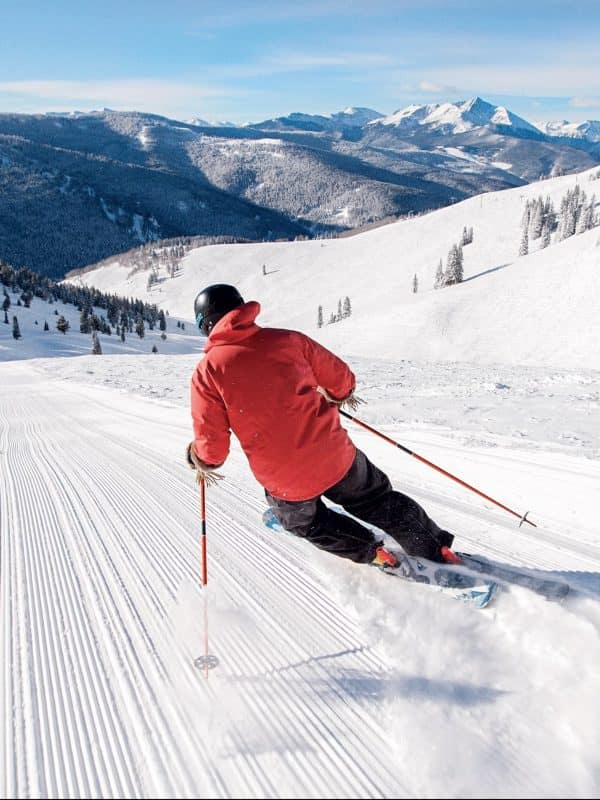 Skier in red jacket carving down pristine groomed slopes with dramatic snow-capped mountain peaks in background